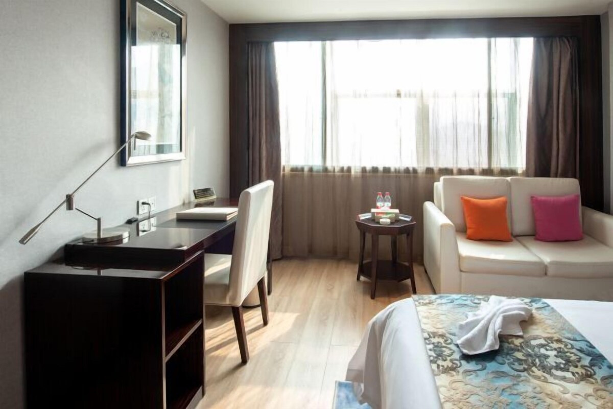 Habitación de hotel con cama, escritorio, silla, una mesita con lámpara y un sofá con cojines.