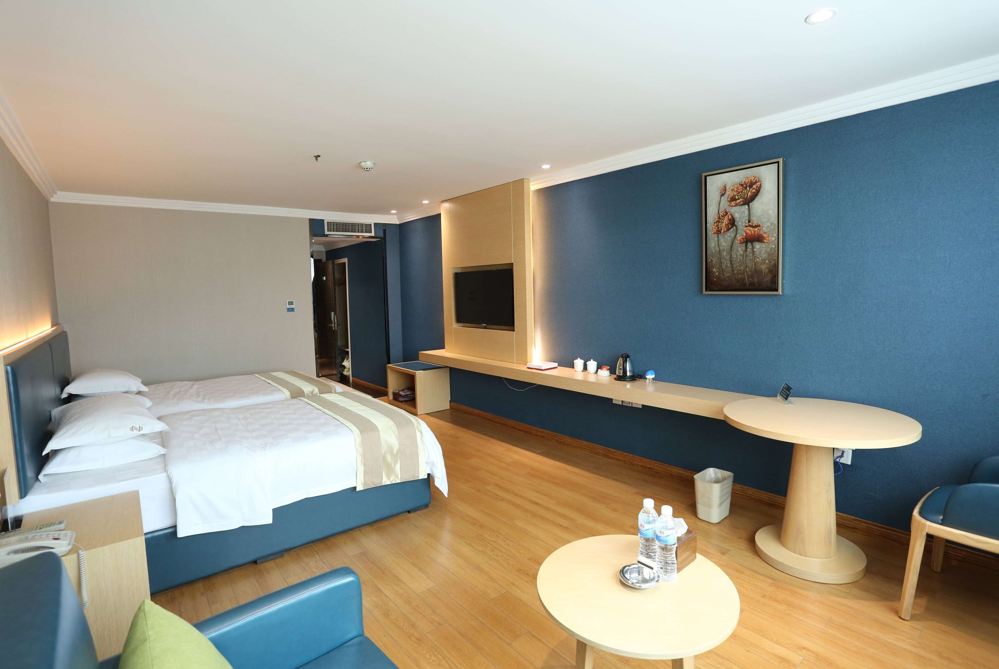 A hotel room with a bed, a desk, a chair, a round table with water bottles, a TV, and a painting on the wall.