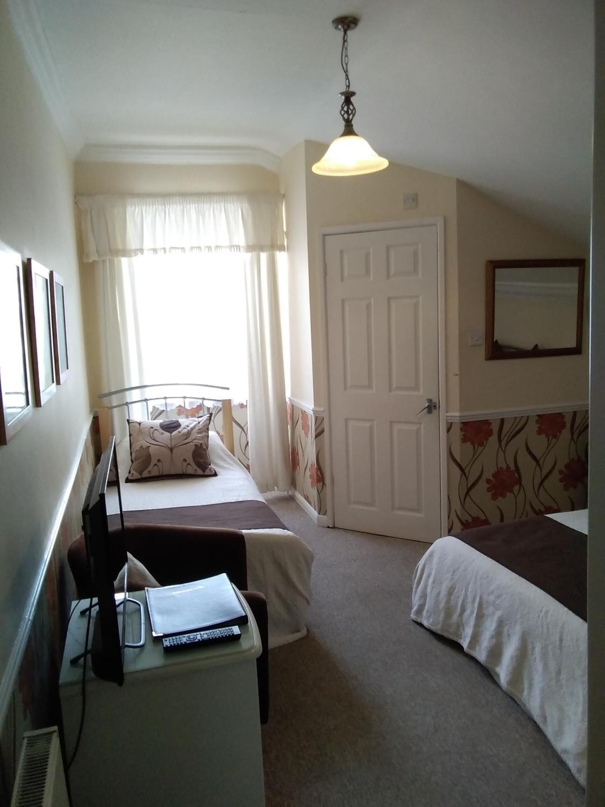 A bedroom with a bed, a desk with a laptop, a chair, a door, and a window with curtains.