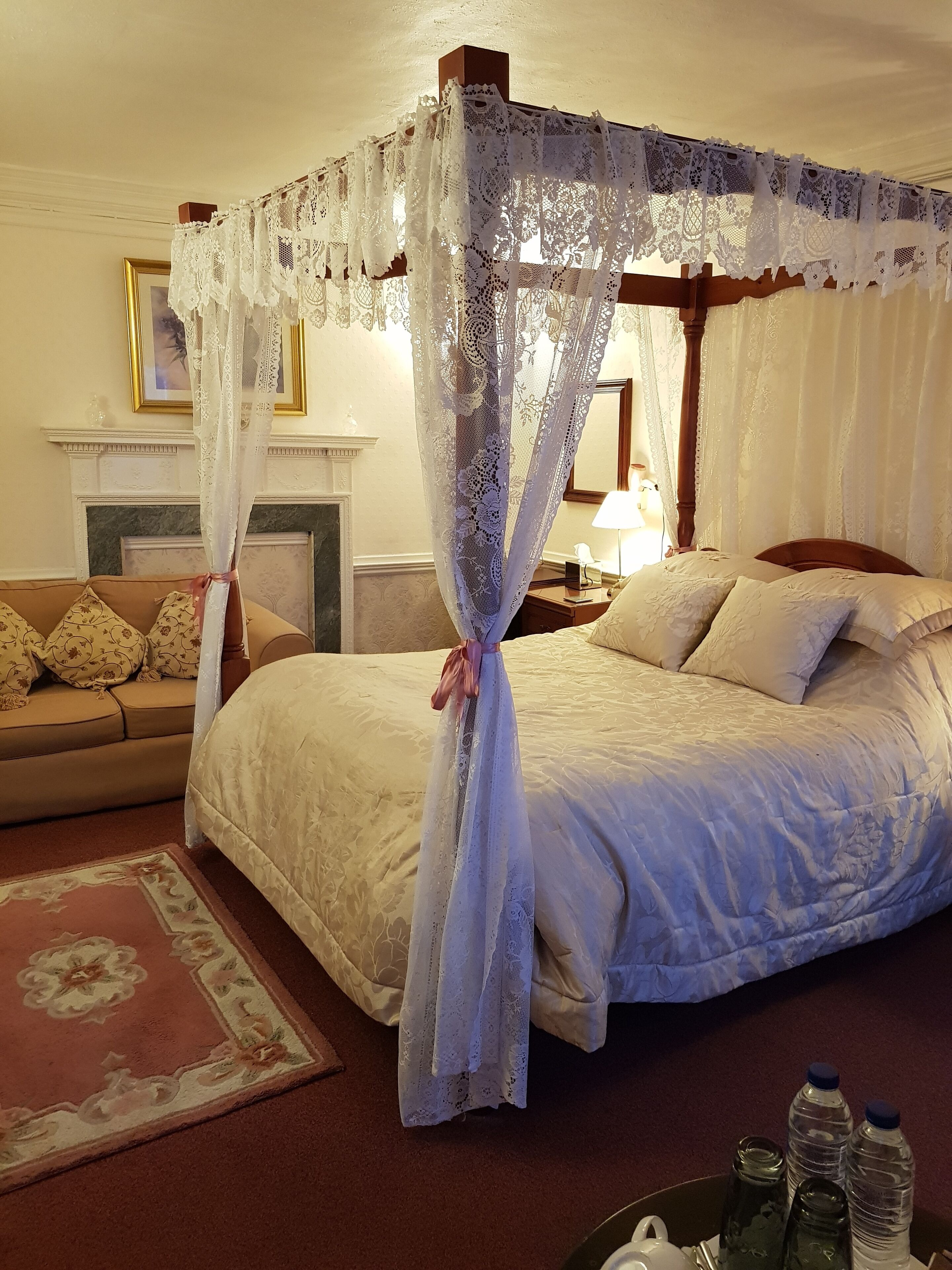 Un dormitorio con cama con dosel, un sofá con cojines florales, una chimenea y una mesita de noche con lámpara.
