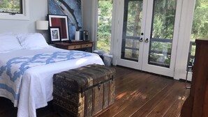 Een slaapkamer met een groot bed, een houten bankje, een schilderij en een glazen schuifdeur naar een balkon.