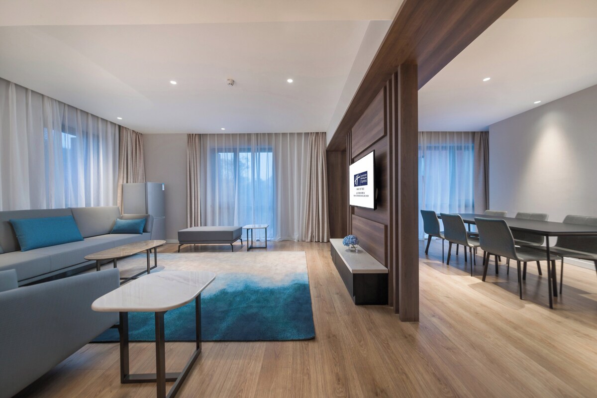 Una habitación de hotel moderna con sofá, una pequeña mesa, un televisor montado en la pared, un rincón de comedor con sillas y un gran ventanal con cortinas.