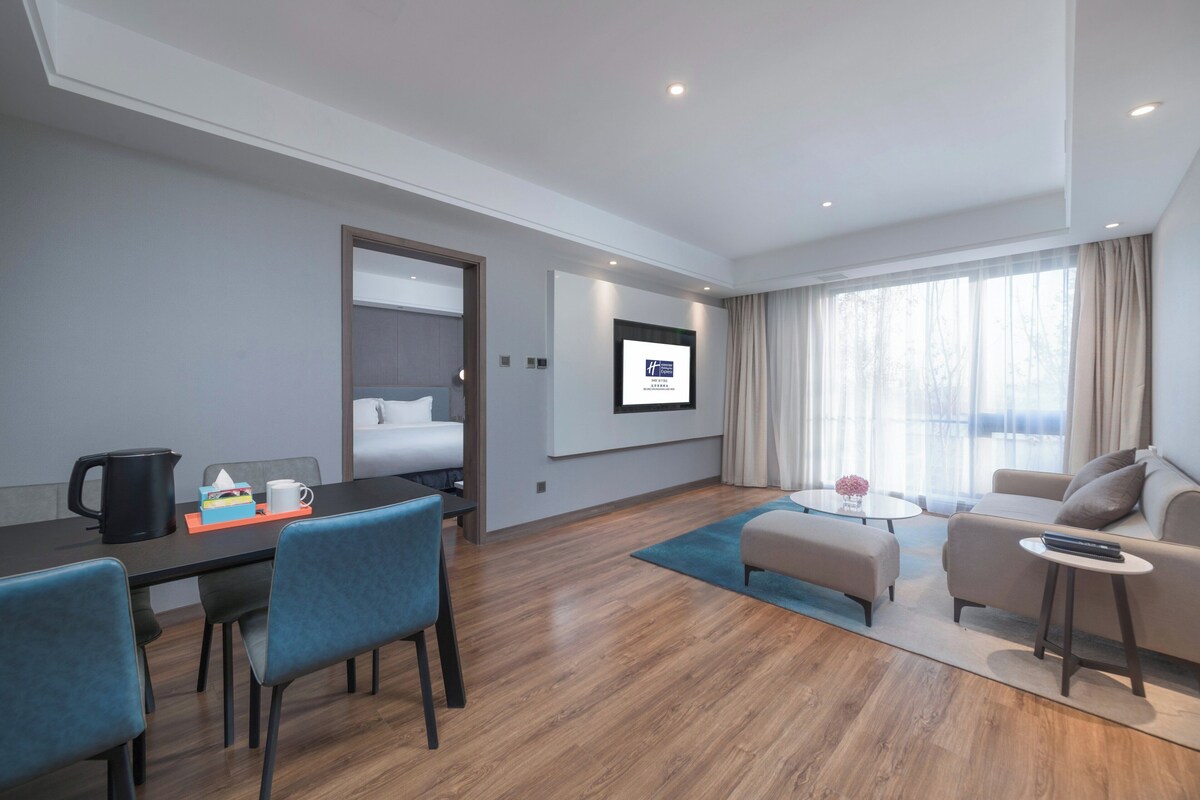 Una habitación de hotel moderna con suelo de madera, una zona de comedor con sillas azules, un sofá y un televisor montado en la pared.