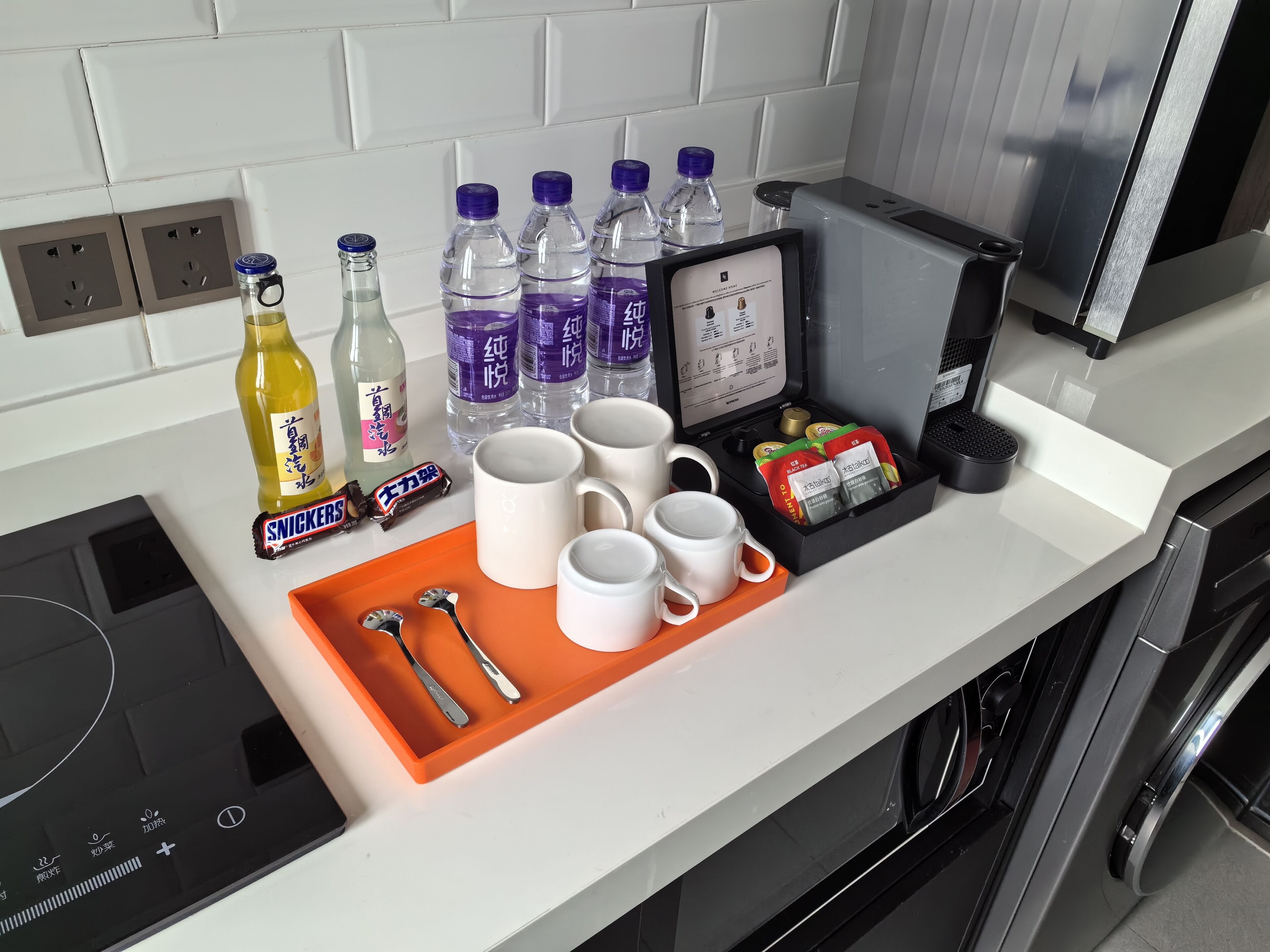 A kitchen counter with a tray containing mugs, a bottle of water, a bottle of juice, a pack of Snickers, a pack of tea, and a coffee machine.