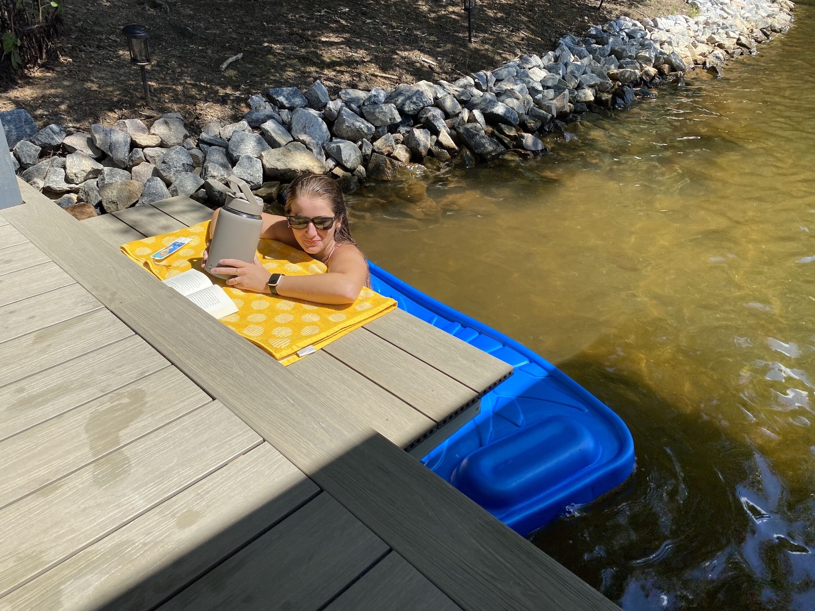 A person relaxing on a wooden deck with a yellow towel and a book, overlooking a river with a rocky bank.