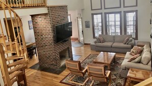 A spacious living room with a brick fireplace, wooden staircase, and a large window with multiple panes. - Spacious Lakefront Home, Amazing View, Great Dock, Sleeps 18 comfortably-Private (Bracey)