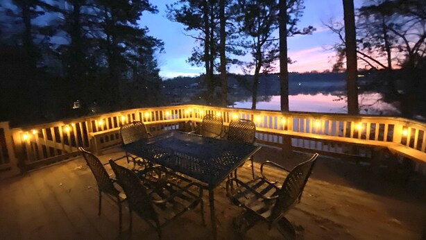 An outdoor patio with a table and chairs, surrounded by a railing with lights, overlooking a body of water and trees. - Spacious Lakefront Home, Amazing View, Great Dock, Sleeps 18 comfortably-Private (Bracey)