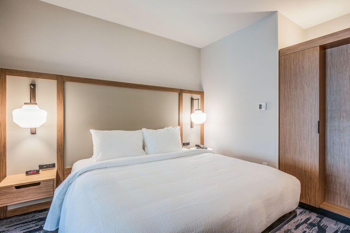 A neatly made bed with white linens, a wooden headboard, bedside lamps, a nightstand, and a wooden wardrobe.