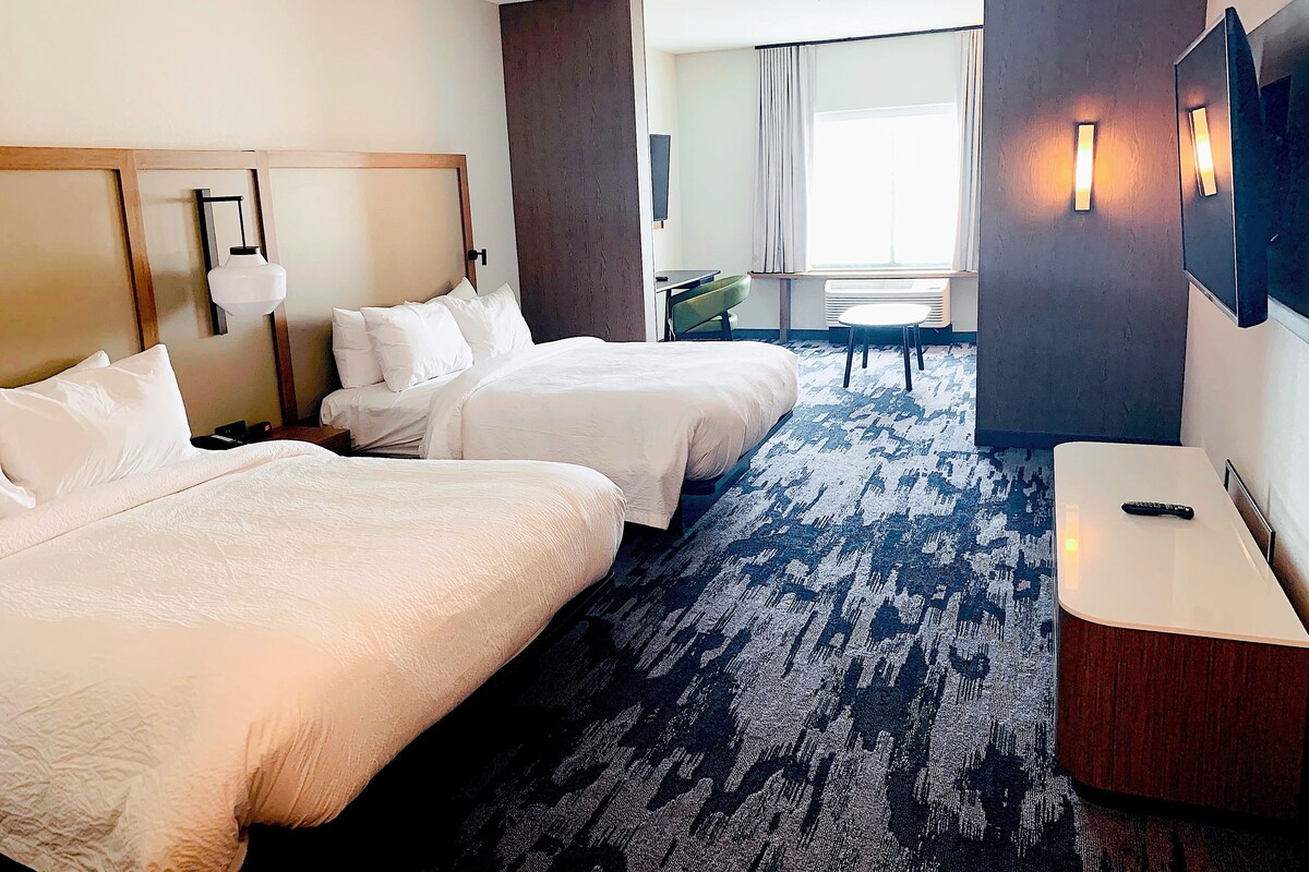 A hotel room with two beds, a desk, a chair, a TV, and a large window with curtains.