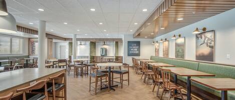 A modern restaurant with wooden tables and chairs, a green cushioned bench, and a menu board on the wall.