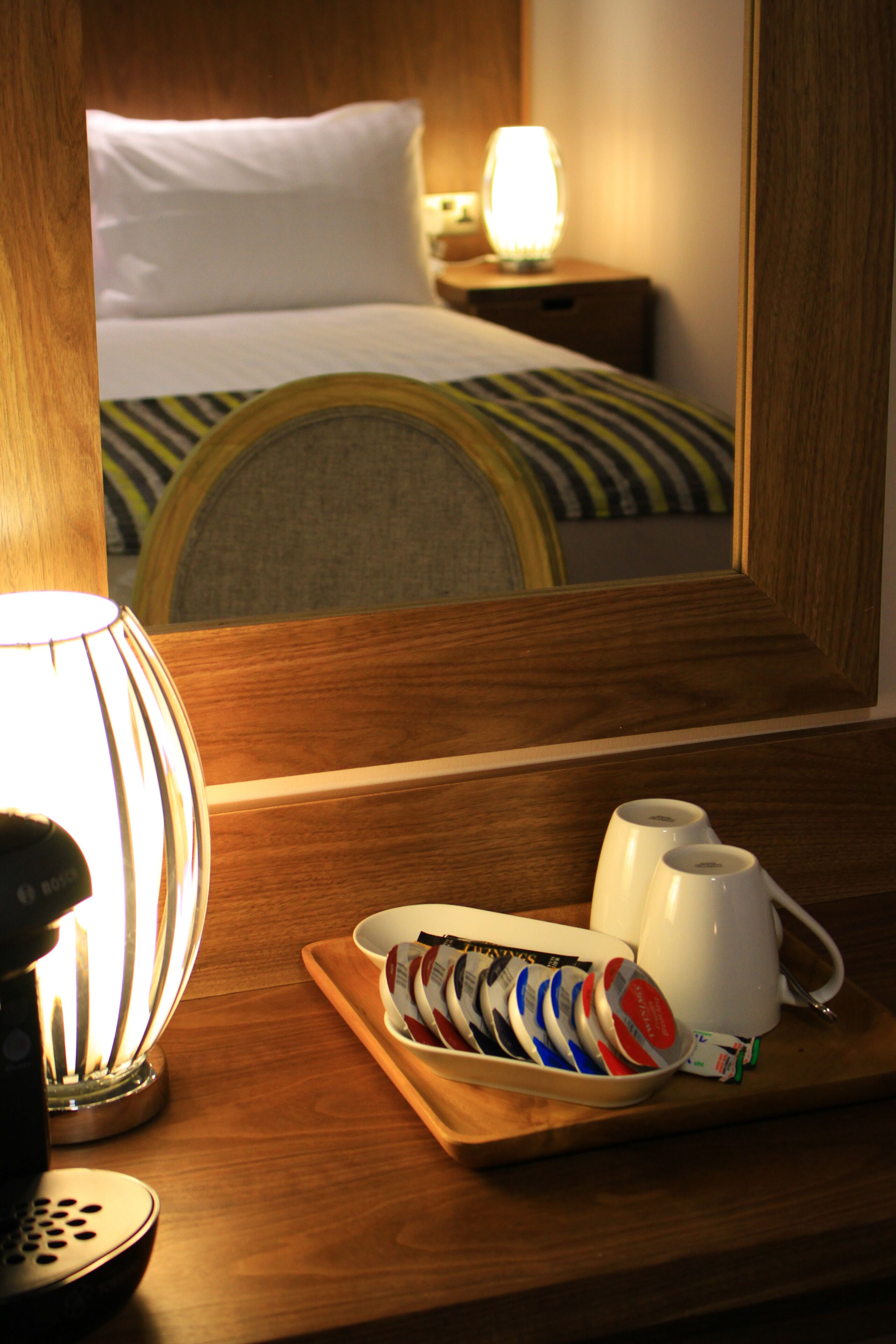 Una habitación de hotel con una mesita de noche de madera, una bandeja con bolsitas de té y una tetera, y una cama con ropa blanca y un almohadón a rayas.