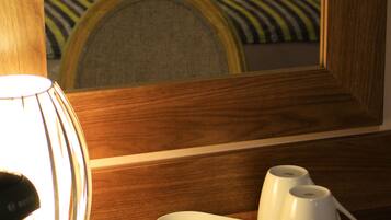 Una habitación de hotel con una mesita de noche de madera, una bandeja con bolsitas de té y una tetera, y una cama con ropa blanca y un almohadón a rayas.