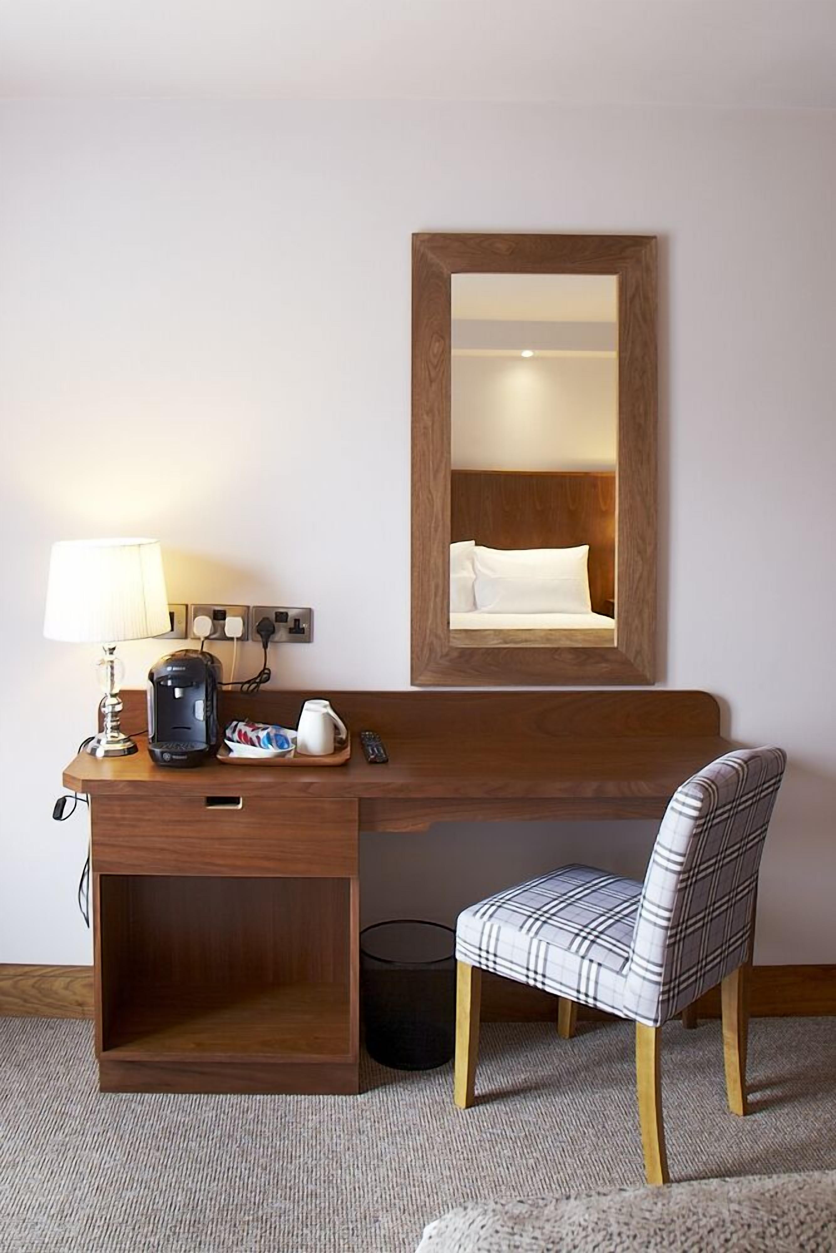 A hotel room with a wooden desk, a lamp, a coffee maker, a remote control, and a plaid chair.