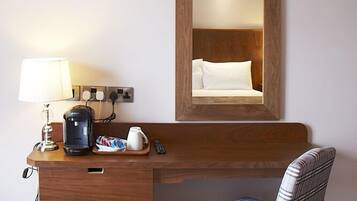 A hotel room with a wooden desk, a lamp, a coffee maker, a remote control, and a plaid chair.