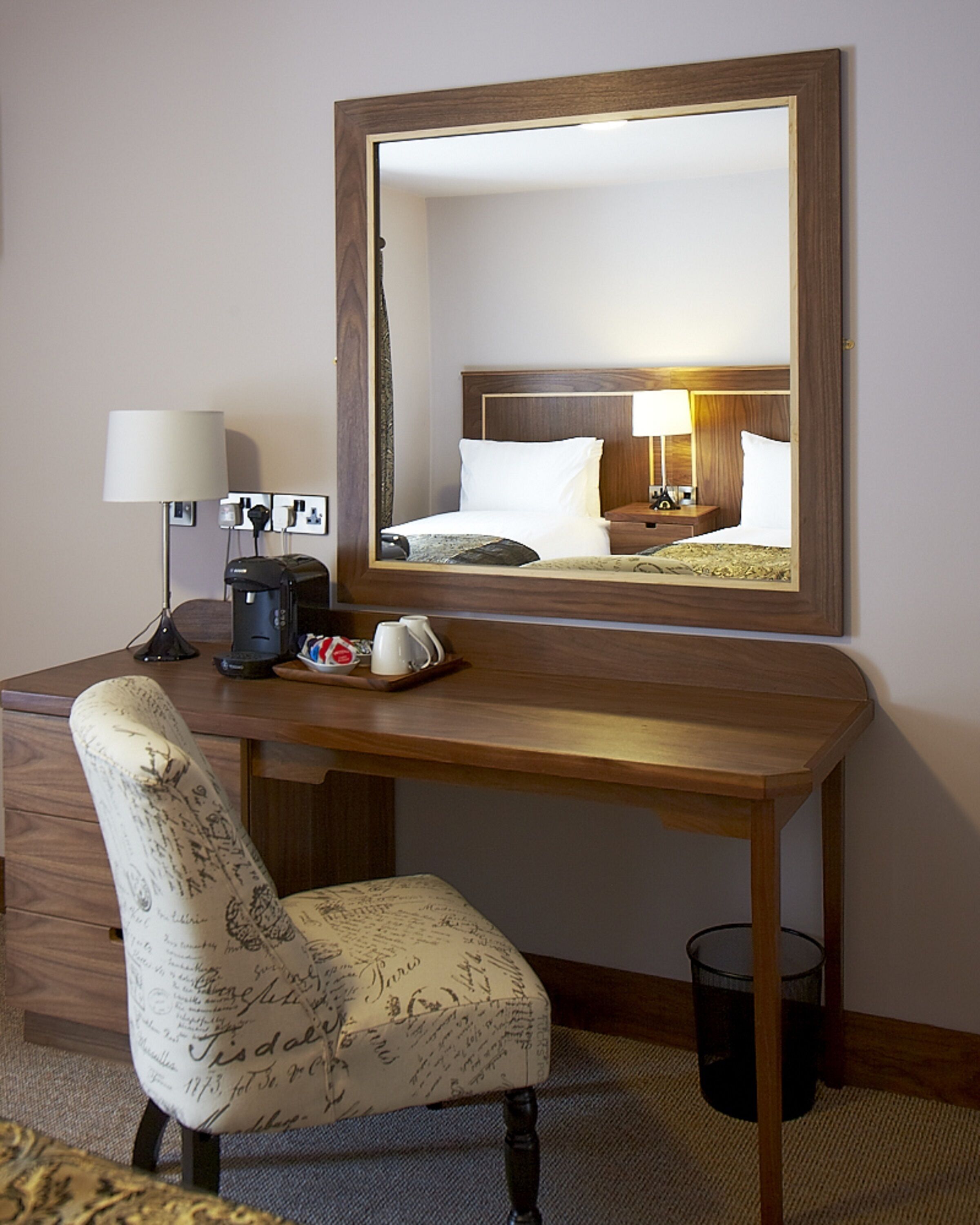 A hotel room with a wooden desk, a chair with a vintage design, a coffee maker, a lamp, and a large mirror reflecting the room.