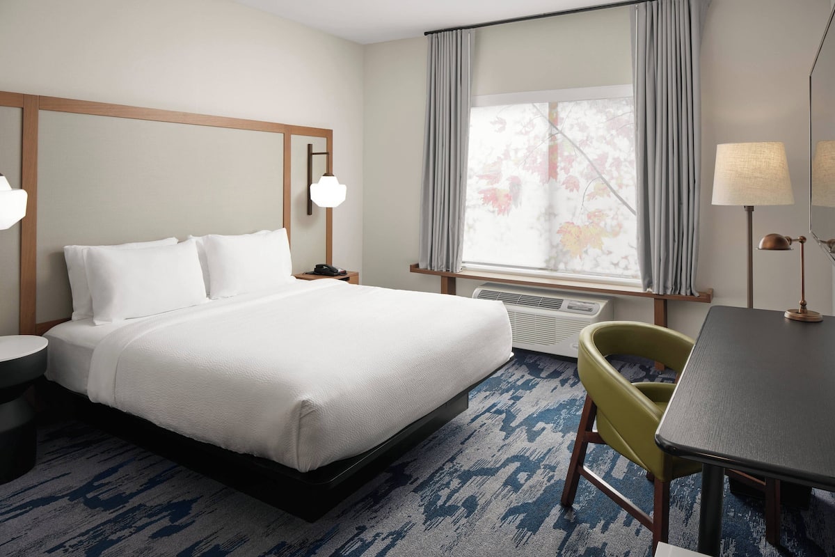 A hotel room with a bed, a desk, a chair, a lamp, and a window with curtains.