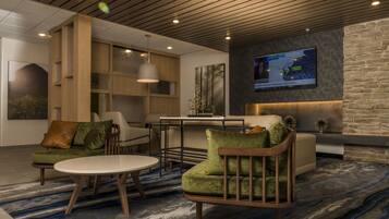 A modern lobby with a wooden ceiling, a large TV screen displaying weather information, and a seating area with green upholstered chairs and a round table.