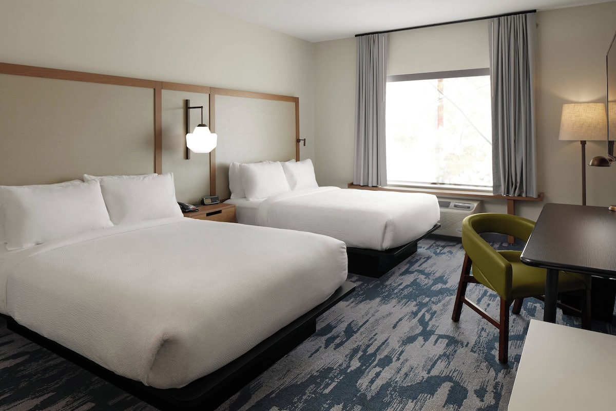 A hotel room with two beds, a desk, a chair, a lamp, and a window with curtains.