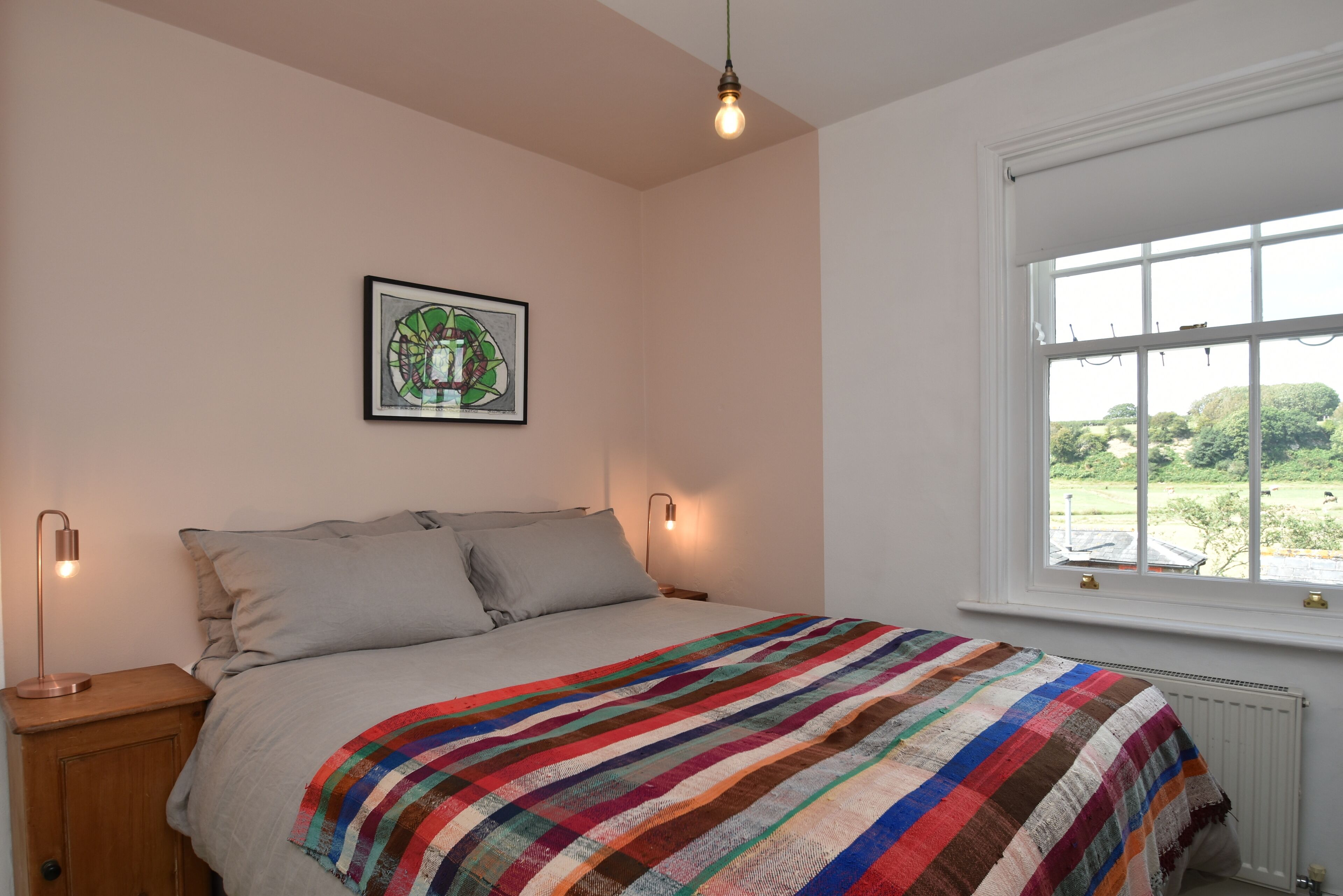 Une chambre à coucher avec un lit, une couverture rayée aux couleurs vives, un tableau encadré accroché au mur et une fenêtre donnant sur un paysage verdoyant.