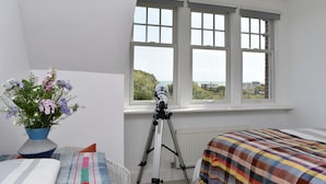 Et soveværelse med et teleskop, en seng med et farverigt tæppe, et lille bord med en vase blomster og et vindue med udsigt over grønne områder og bygninger.