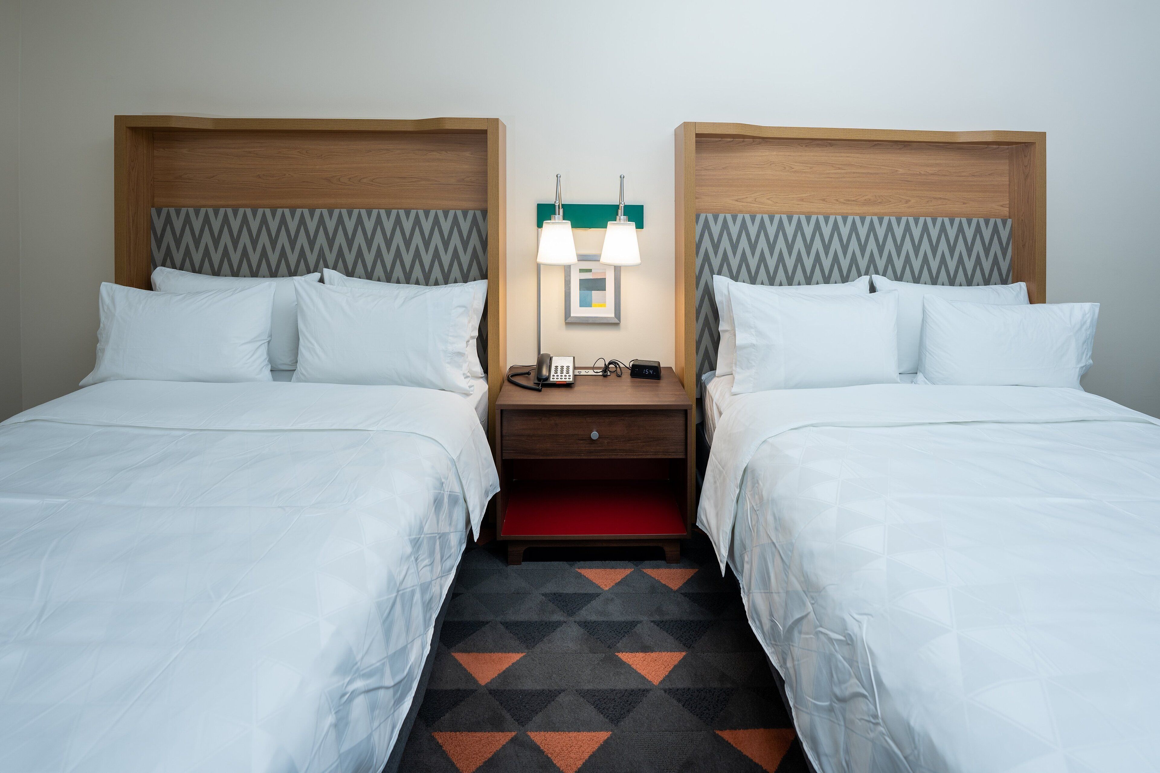 A hotel room with two beds, a nightstand with a phone and lamp, and a patterned carpet.