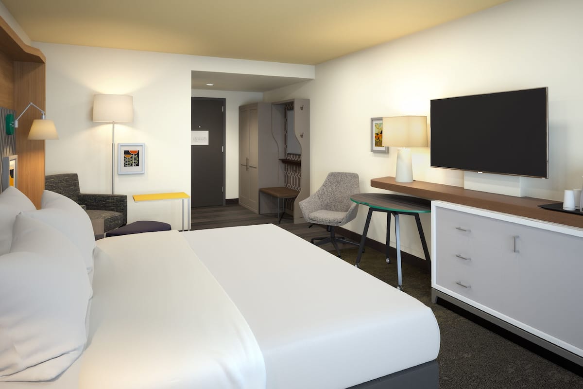 Una habitación de hotel moderna con una cama grande, un escritorio con televisor de pantalla plana, una silla y una mesita.