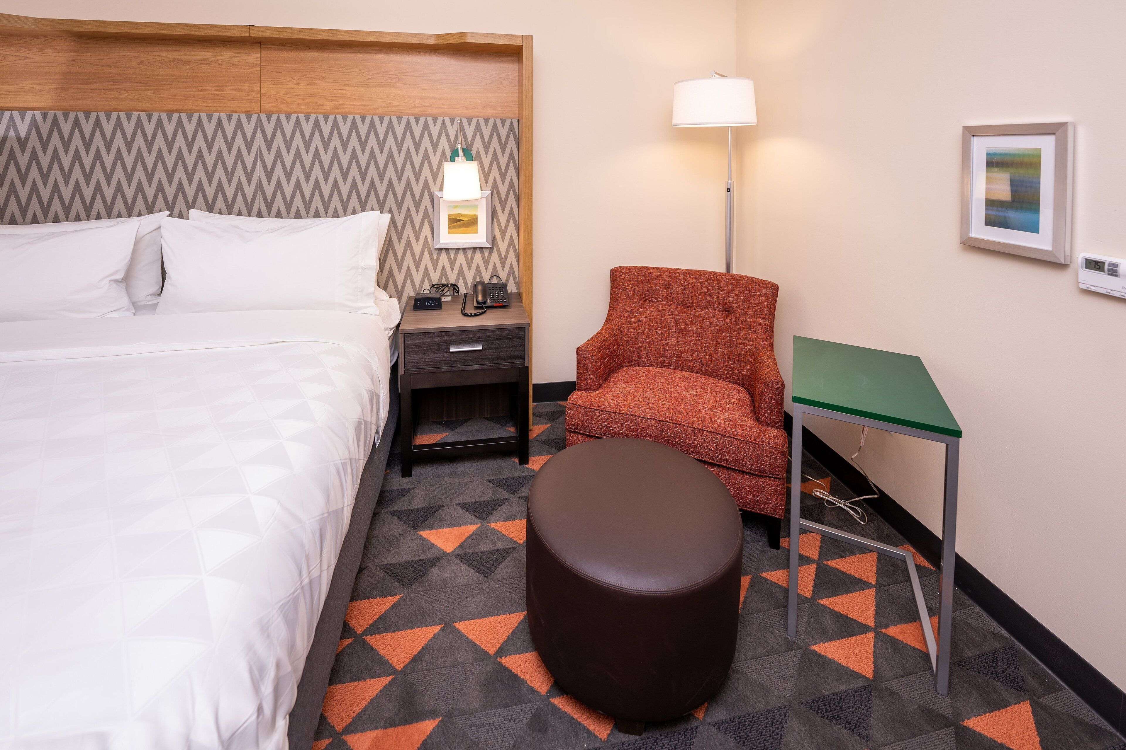 Une chambre d’hôtel avec un lit, une table de chevet, une lampe, une chaise, une table et un tapis à motifs.
