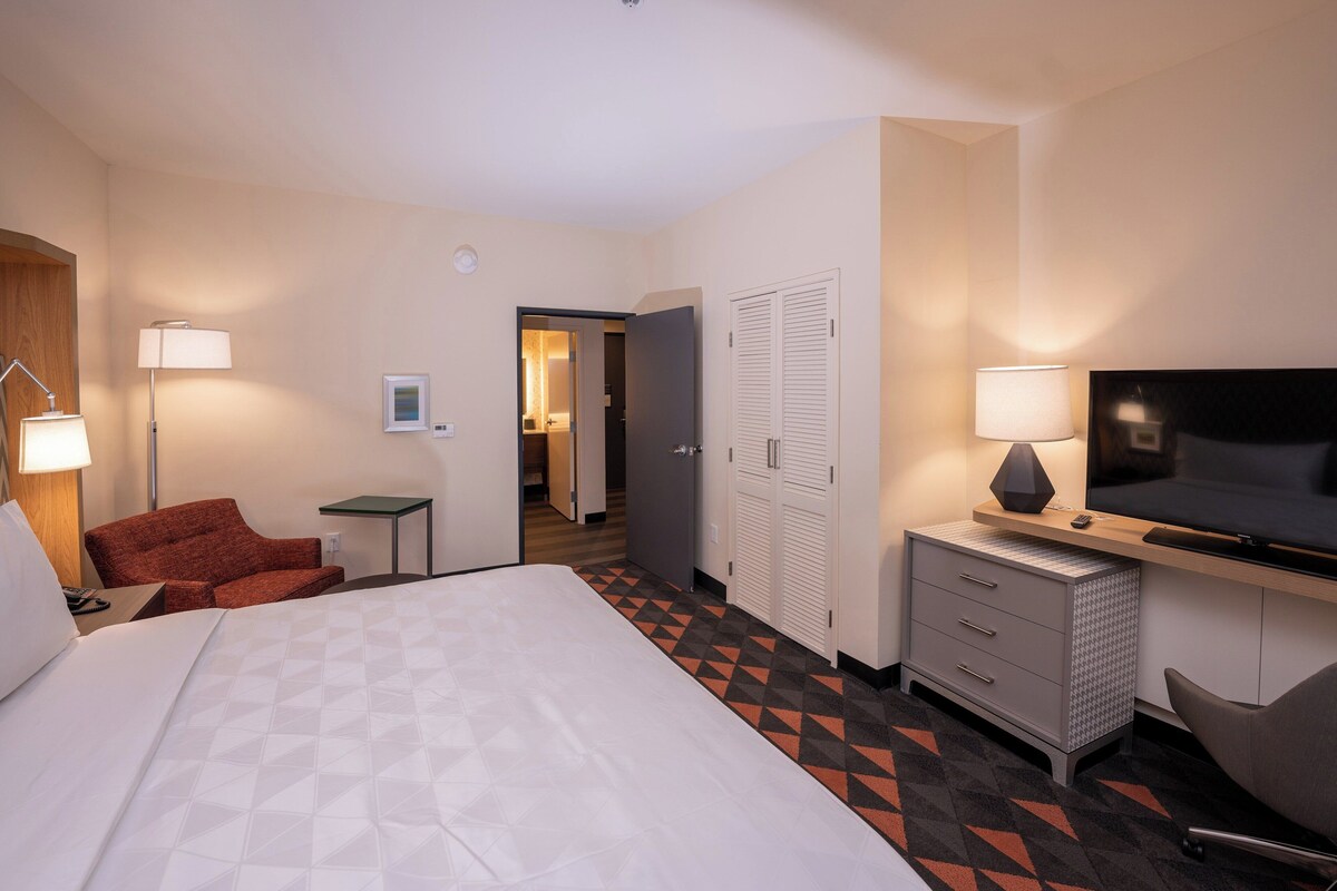 A hotel room with a bed, a desk with a TV, a chair, a lamp, and a wardrobe.