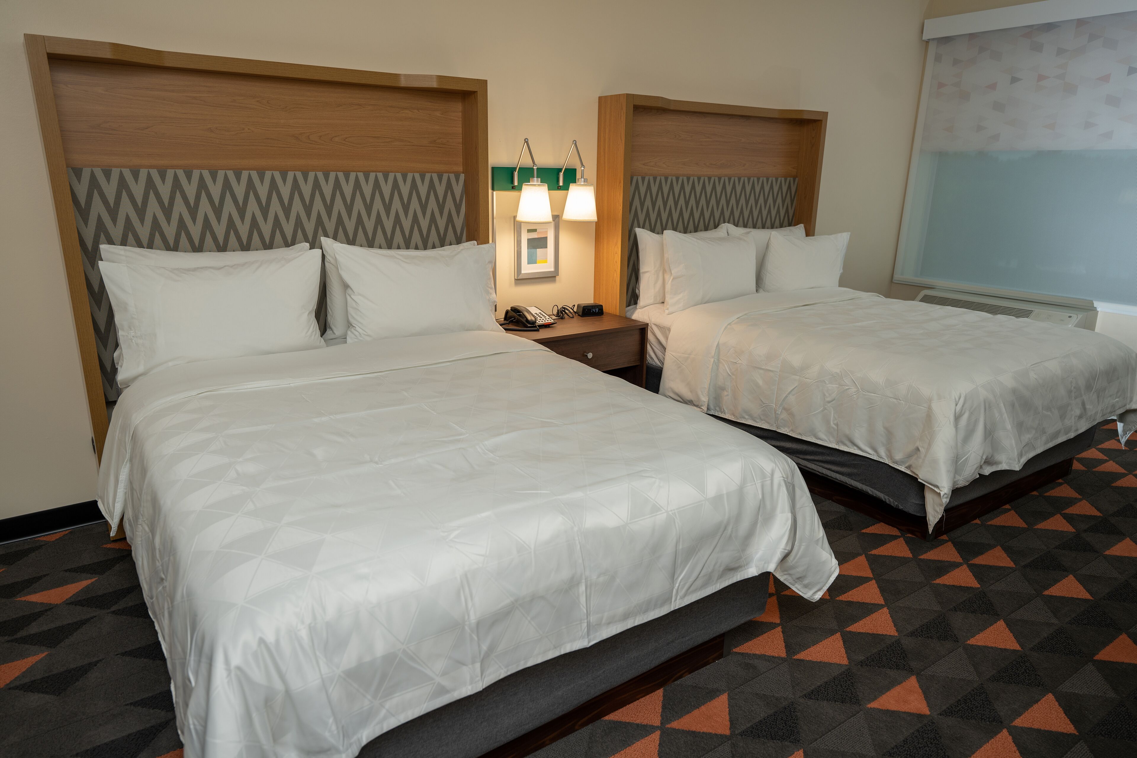 A hotel room with two beds, a nightstand with a phone and lamp, and a window with a view.