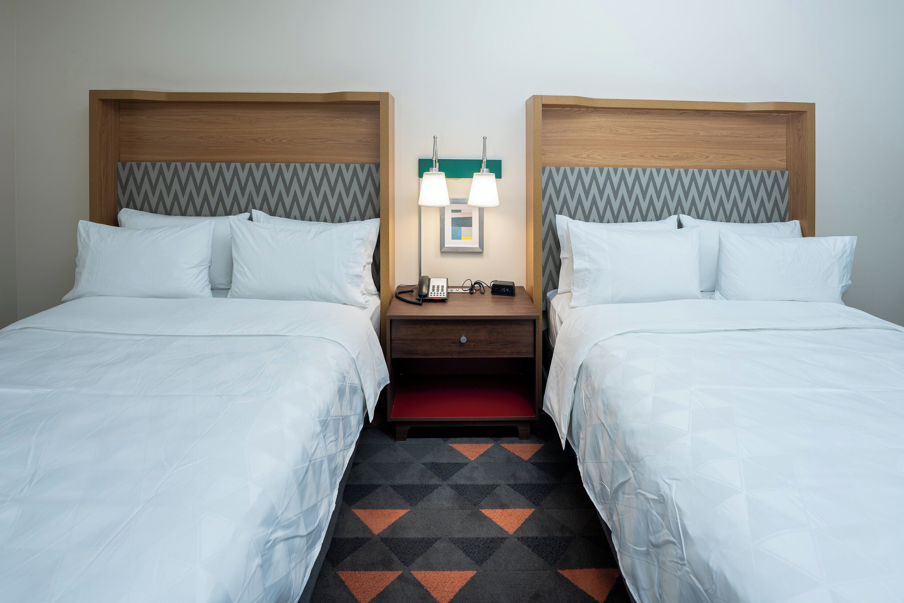 Une chambre d’hôtel avec deux lits, une table de chevet avec un téléphone et une lampe, et une moquette à motifs.