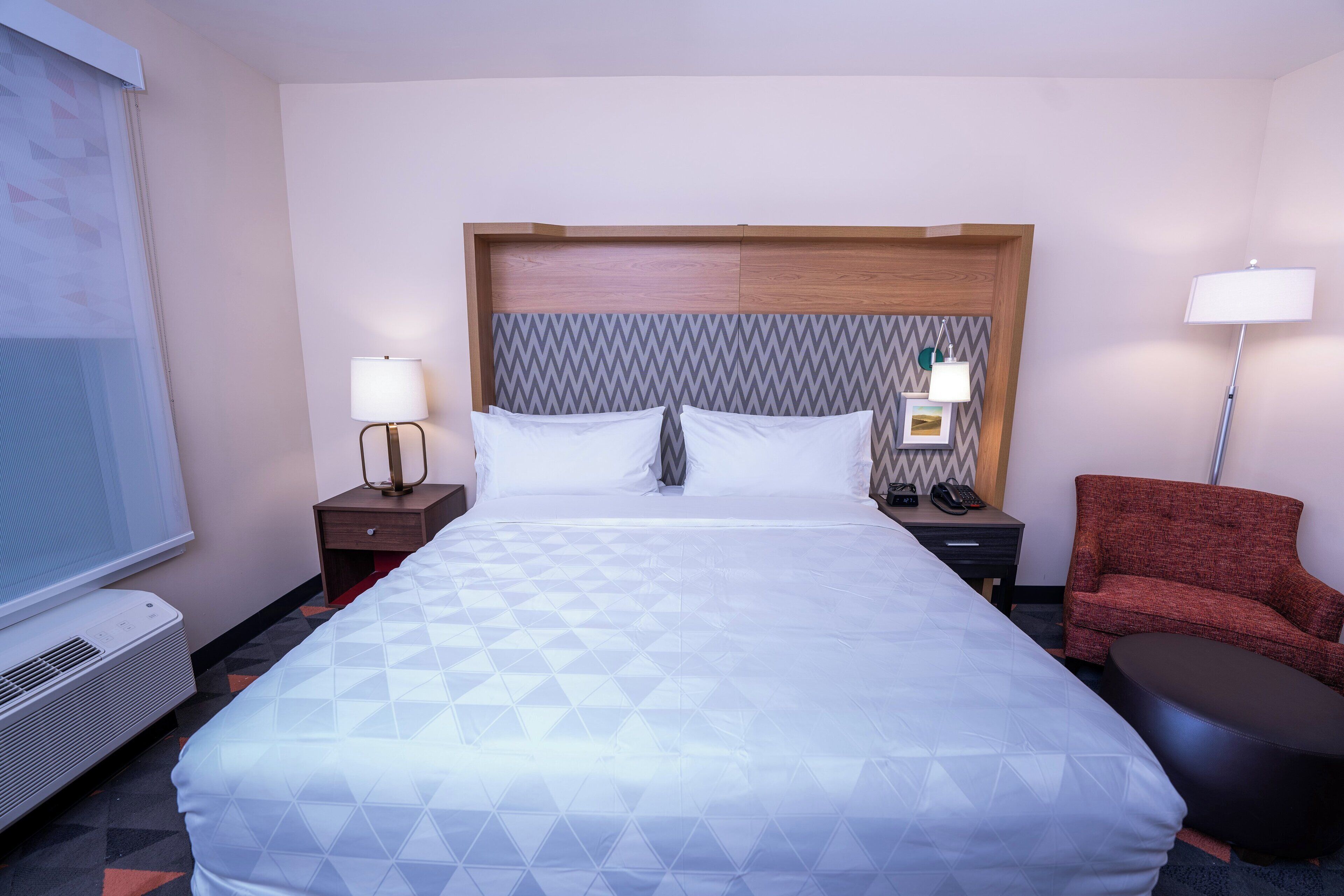 Una habitación de hotel con una cama grande, dos mesitas de noche con lámparas, un sillón rojo, una mesita pequeña y una pantalla para proyector.