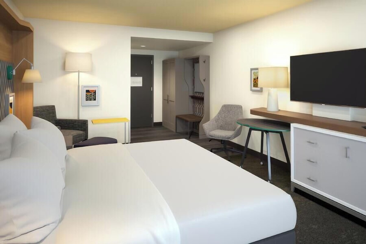 A hotel room with a large bed, a desk with a flat-screen TV, a chair, and a small table.