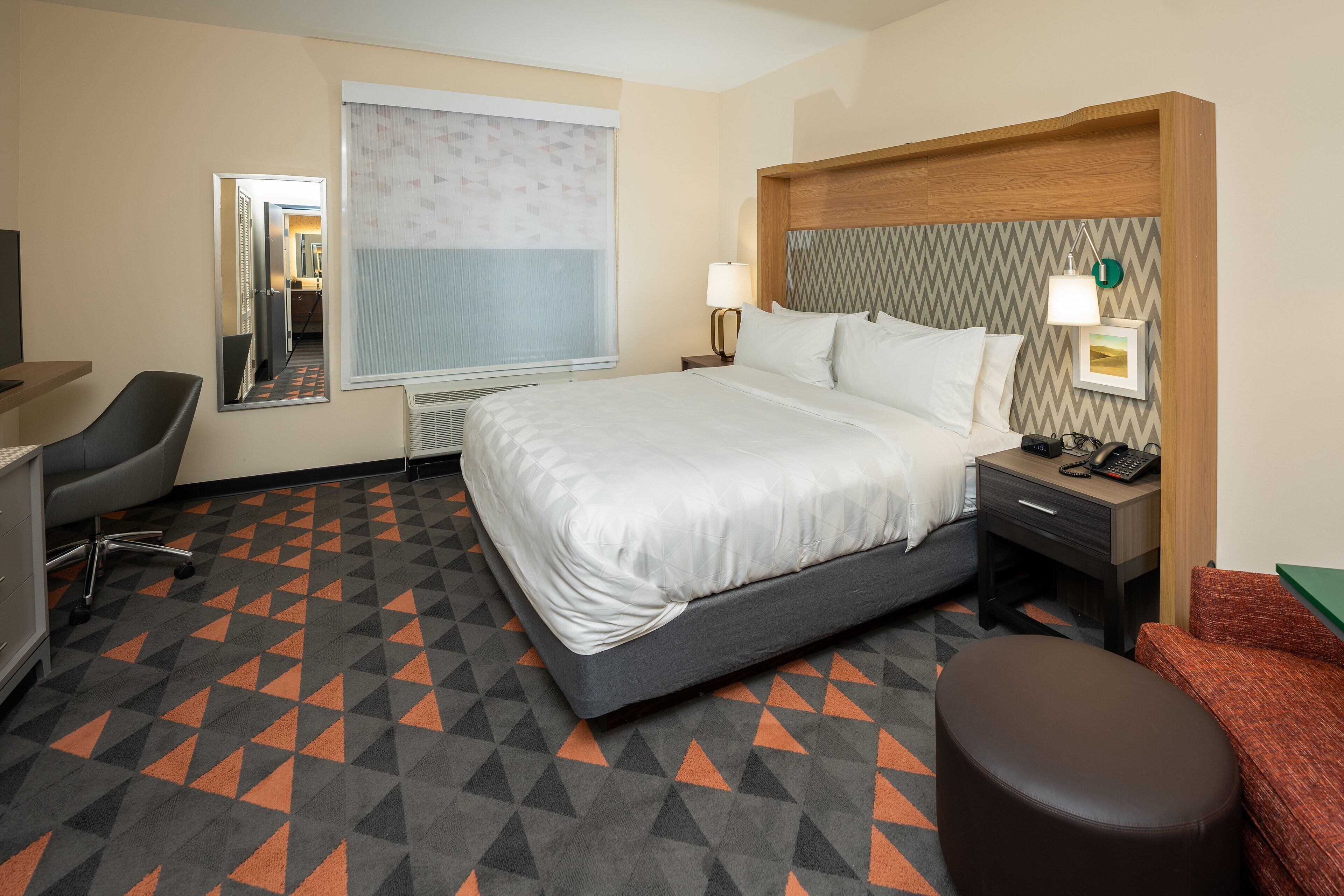 Ein Hotelzimmer mit Bett, Schreibtisch, Stuhl, Nachttisch, Lampe und einem Fenster mit Jalousien.
