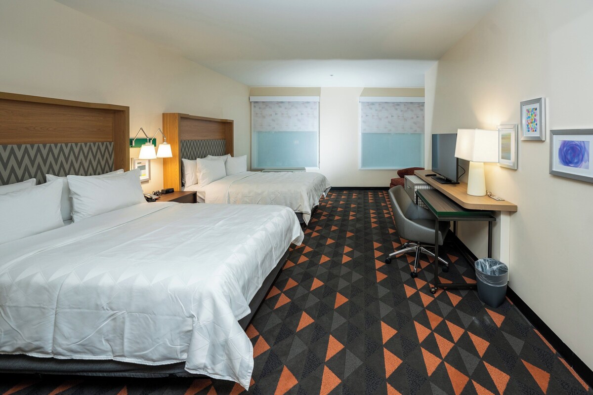 A hotel room with two beds, a desk, a chair, a trash bin, and a window with blinds.