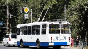 Un autobús blanco y azul con el número 1828 en la parte trasera, circulando por una carretera con una señal de paso de peatones y un semáforo al fondo.