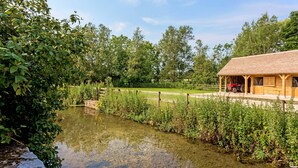A wooden cabin with a porch, surrounded by lush greenery and a calm pond.
