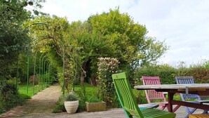 Een houten terras met een tafel en kleurrijke stoelen, omringd door weelderige begroeiing en een tuinpad.
