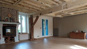 Een ruime kamer met houten vloer, open haard, groot raam met luiken en een rustiek balkenplafond.