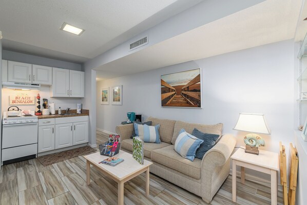 A modern living room with a beige sofa, a coffee table, and a kitchen area with white cabinets and a stove. - Hip Nautic - Couple's Getaway, Indoor Pool Hot Tub, Walk to Beach, Pickleball (Hilton Head Island)