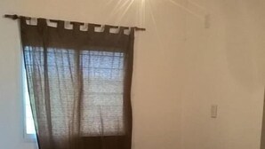 Une petite pièce faiblement éclairée, avec un lit simple, une fenêtre ornée de rideaux légers et un luminaire au plafond.