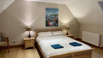 A bedroom with a sloped ceiling, a wooden bed, two bedside lamps, a nightstand, a chair, and a painting of a coastal scene.