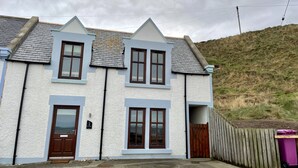 A two-story house with a white exterior and dark brown trim, featuring a front door, two windows on the ground floor, and a small porch area. - Modern Fisherman's Cottage with Stunning Uninterrupted Views of the Moray Firth (Findochty)