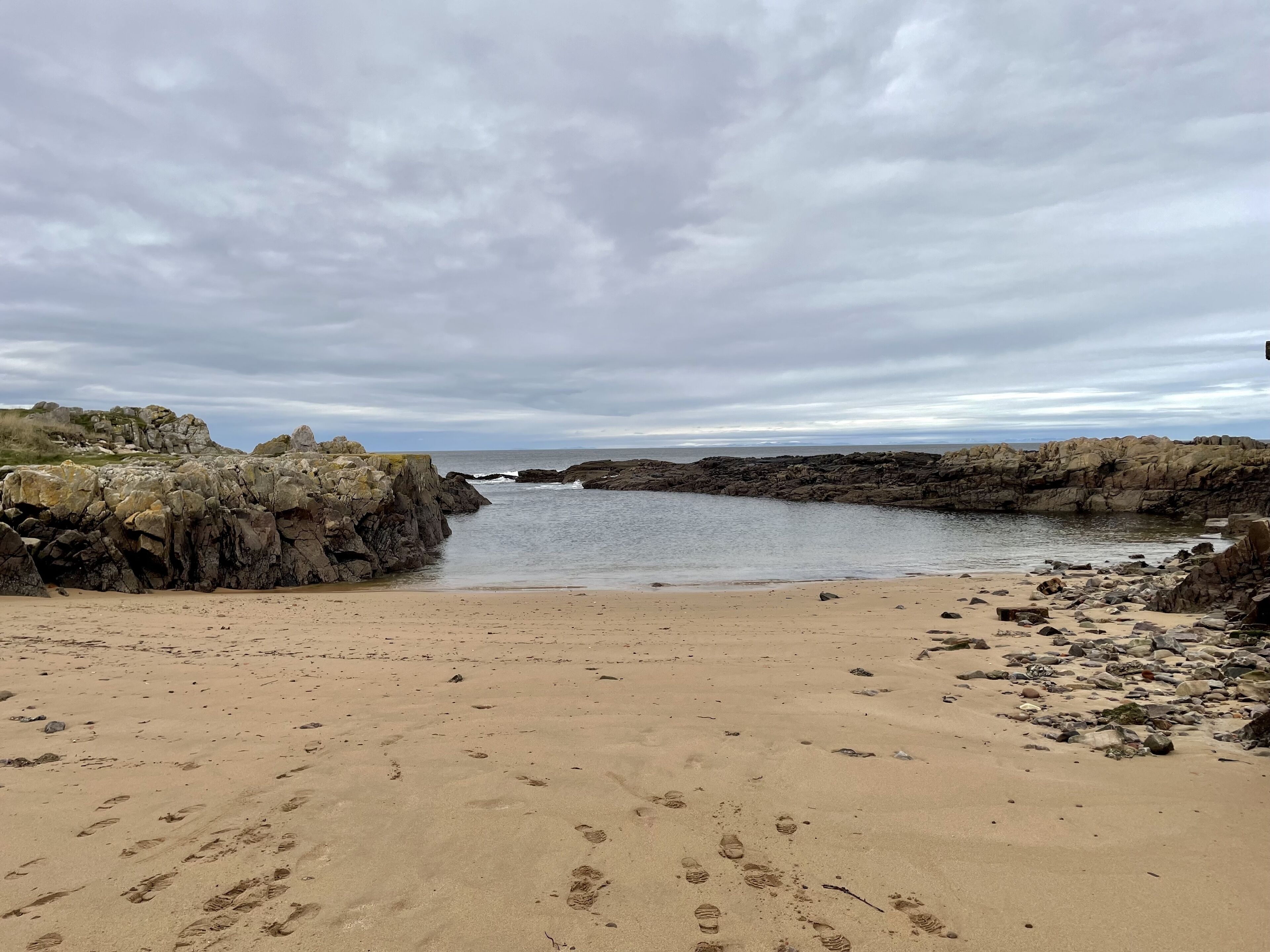 Une plage rocheuse, parsemée d’empreintes dans le sable, sous un ciel nuageux.