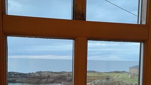 A view through a window of a coastal area with a sandy beach, rocky cliffs, and a grassy area with benches. - Modern Fisherman's Cottage with Stunning Uninterrupted Views of the Moray Firth (Findochty)