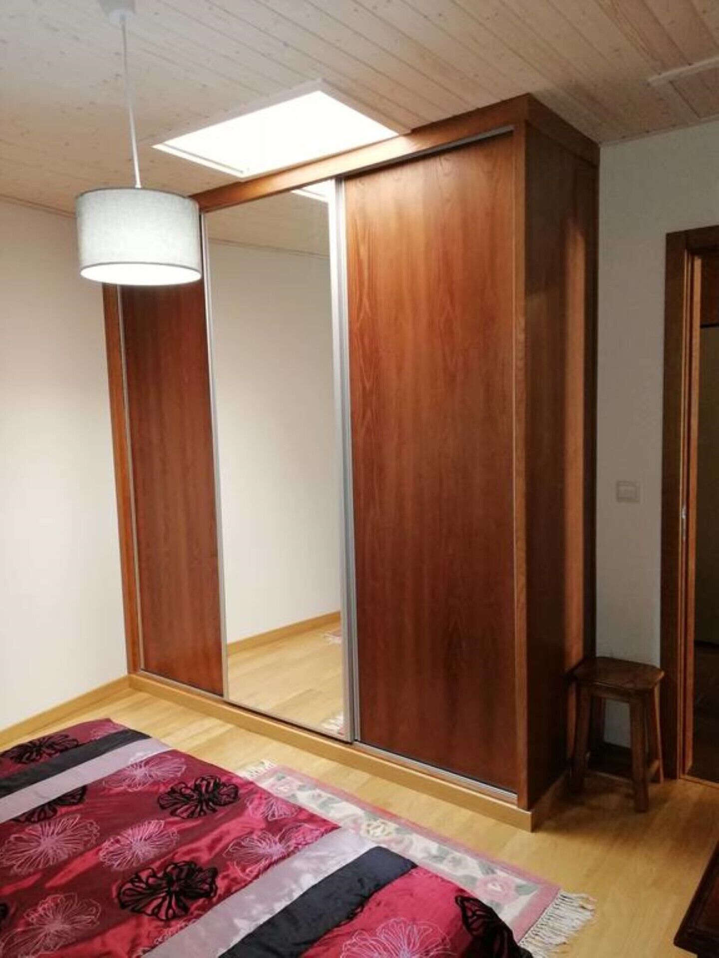A bedroom with a wooden wardrobe, a mirror, a hanging light fixture, and a patterned bedspread.