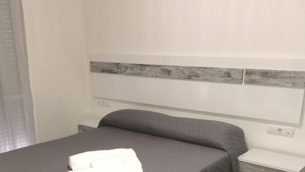 A neatly made bed with a gray quilt and white towels, a wooden headboard, a bedside table, and a nightstand with a lamp. - Mariñiguez Apartment. (Jerez de la Frontera)