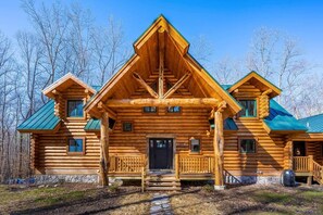 Una baita di legno con un camino centrale, un portico con ringhiere e fondamenta in pietra.
