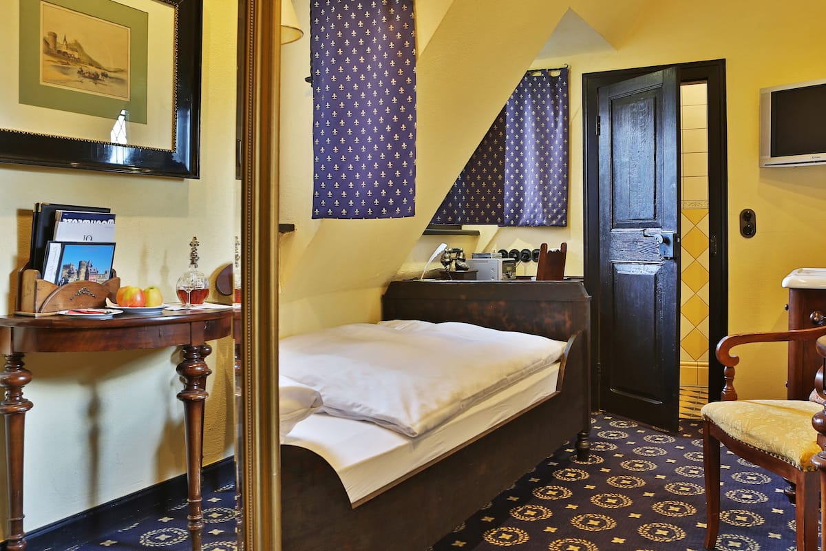 A hotel room with a bed, a desk with a computer, a chair, a small table with apples and a glass, and a bathroom visible through an open door.