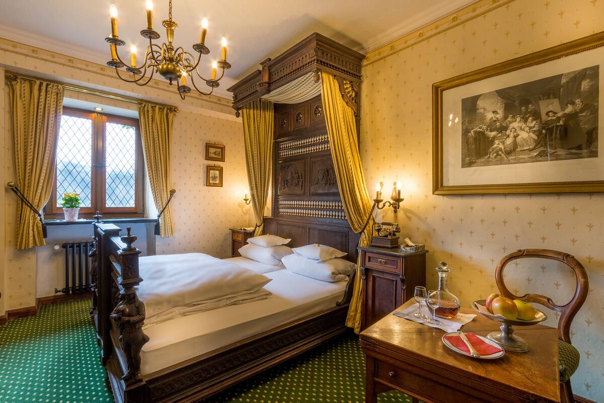 A classic room with a four-poster bed, a wooden desk with a mirror, a framed picture on the wall, and a window with curtains.