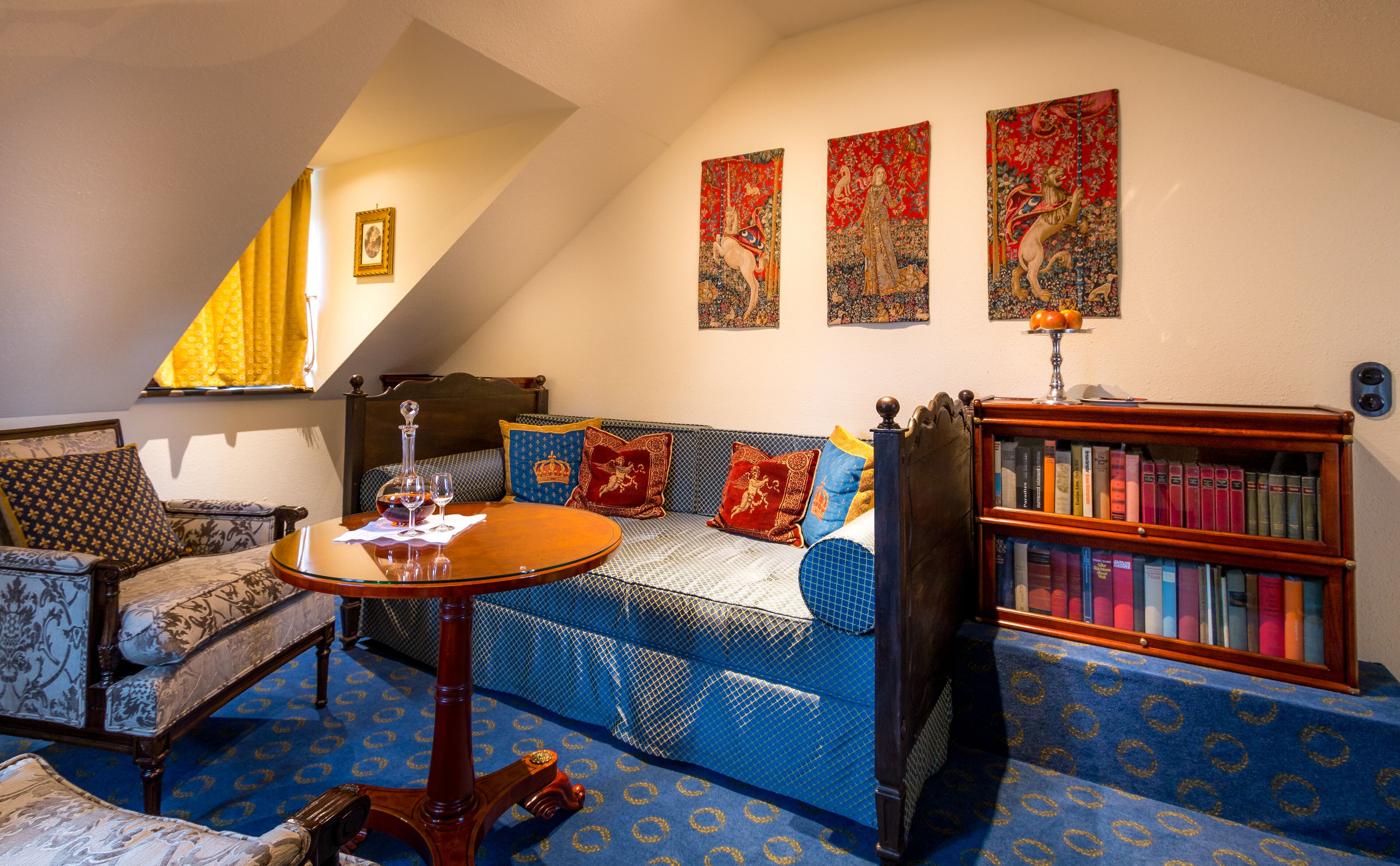 A cozy living room with a blue patterned sofa, a wooden coffee table, a bookshelf, and a window with curtains.